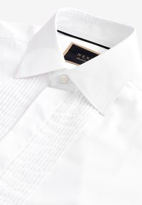 Camicia bianca con colletto classico, parte frontale plissettata, chiusura con un solo bottone e etichetta nera con la scritta "NEX" all'interno del colletto.