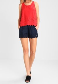 Röd ärmlös topp med laserutskuren vågig nederkant över marinblå shorts med liknande detaljer. Bärs med svarta slide-sandaler.
