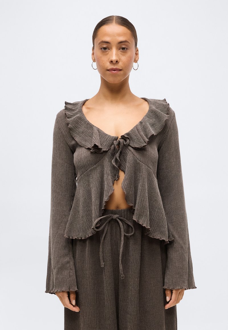 Γυναίκα που φοράει καφέ ριμπ πλεκτό τοπ με βολάν στο γιακά, δέσιμο μπροστά και ταιριαστά παντελόνια με κορδόνι, μπροστά σε απλό φωτεινό φόντο.