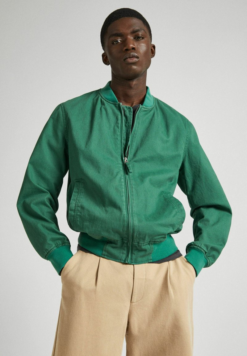 Chaqueta bomber verde con cremallera frontal, puños acanalados y cinturilla. Fabricada con un material suave, presenta bolsillos laterales y un cuello alto.