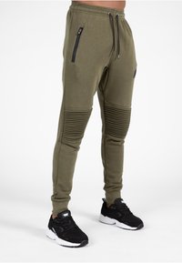 Olijfgroene joggers met ribgebreide kniepanden, ritszakken aan de zijkant en een trekkoord in de taille. Gecombineerd met zwarte hardloopschoenen.