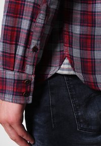 Camicia in flanella a quadri rossa e blu con polsini abbottonati, mostrata su una mano; tessuto denim visibile in vita con una sottile texture.