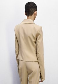 Blazer beige su misura con colletto affilato e spalle strutturate, realizzato in tessuto liscio, caratterizzato da una silhouette sfiancata e cuciture sottili.
