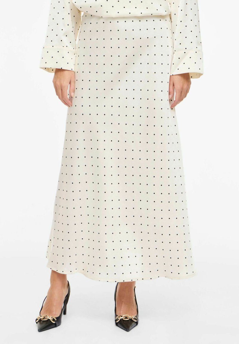 Jupe longue couleur crème à pois noirs, en tissu lisse. Accentuée d'un léger éclat et associée à des talons noirs pointus.