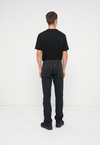 Čierne tričko a tmavomodré džínsy, slim fit s viditeľným šitím a zadnými vreckami, nosené s čiernymi topánkami na jednoduchom pozadí.