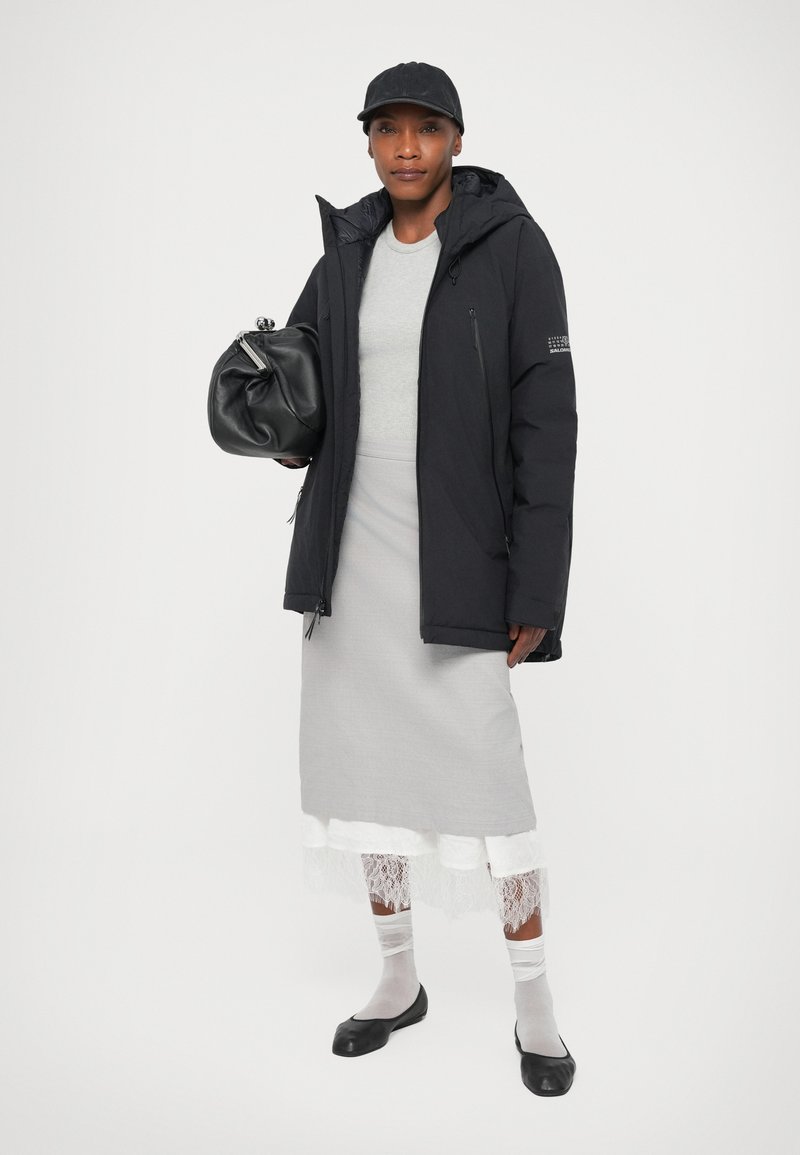 Femme portant une casquette noire, une veste noire, un haut gris, une jupe grise avec une bordure en dentelle blanche, des chaussettes blanches, des ballerines noires, tenant un sac à main noir.