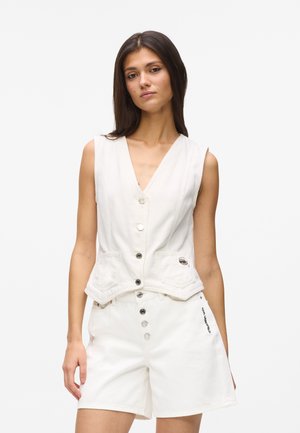 Frau trägt ärmellose weiße Weste mit Knöpfen und weiße Shorts mit hoher Taille und Knopfdetails, stehend vor einfachem hellem Hintergrund.