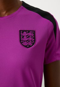 Purple athletic t-shirt with black shoulder accents. Features a textured, breathable fabric and a crest with decorative elements on the chest.