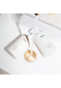 Två OUAI-produkter: en vit flaska med handtvål och en vit lotionflaska, bredvid ett guldavloppslock, med vatten som rinner över dem.