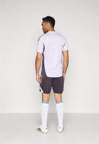 Sivé športové šortky s bielymi akcentmi, doplnené svetlofialovým tričkom s krátkym rukávom. Biele ponožky s čiernymi pruhmi a čierne kopačky dotvárajú vzhľad.