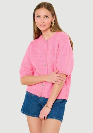 Jonge vrouw die een roze gebreid vest met knopen en korte mouwen draagt, gecombineerd met een denim minirok, afgewerkt met gouden oorbellen en een armband.