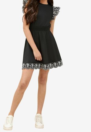 Czarna sukienka z krótkimi, falbaniastymi rękawami z motywem kwiatowym. Spódnica ma zaokrąglony brzeg z misterną białą haftowaną dekoracją. Dopełniona białymi trampkami.