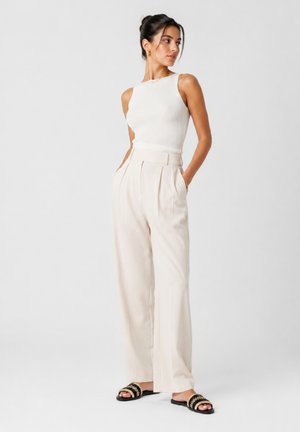 Mujer con camiseta blanca sin mangas, pantalones beige de talle alto y pernera ancha, y sandalias negras con tiras trenzadas, de pie con las manos en los bolsillos.
