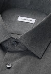 Gros plan sur le col d'une chemise grise à texture fine avec des boutons noirs et une étiquette blanche cousue portant l'inscription « seidensticker ».