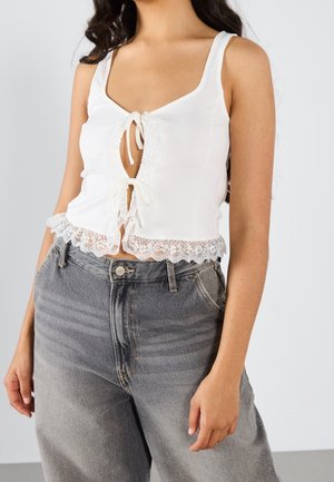 Kvinde iført en hvid ærmeløs crop top med blondekant og bindebånd foran, kombineret med højtaljede, falmede grå jeans.