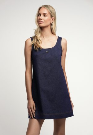 Blondynka w ciemnoniebieskiej, bezrękawnikowej mini sukience z denimu, stoi z jedną ręką opuszczoną, patrząc w lewo na prostym jasnym tle.