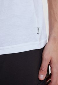 Hvid bomulds t-shirt med en rund kant, der har et synligt mærke på siden. Hånd hviler mod mørkt stof.