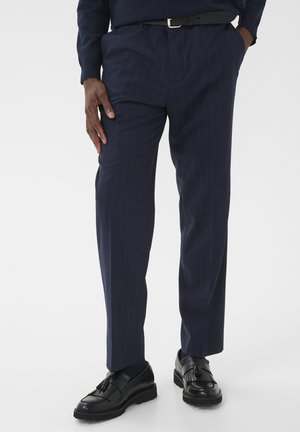 Hombre con pantalones azul marino de rayas, cinturón negro y mocasines de cuero negro con flecos, de pie con una mano en el bolsillo y la otra al lado.