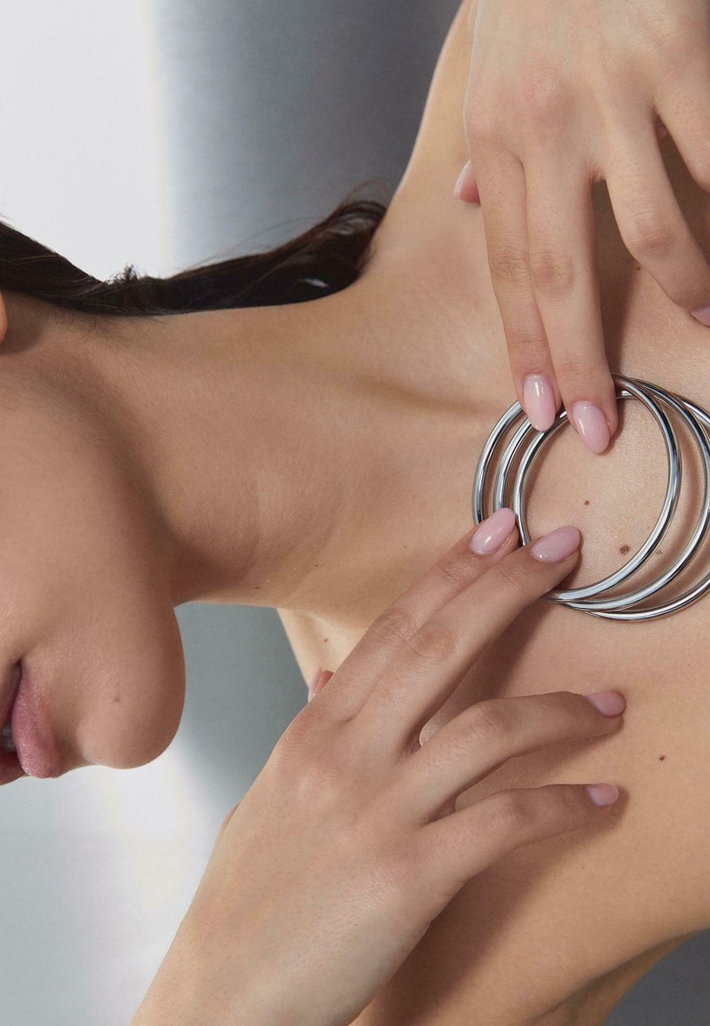 Femme aux ongles rose clair tenant trois bracelets argentés près de sa clavicule sur un fond neutre.