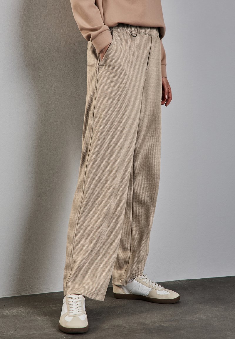 Pantaloni beige a gamba larga in un tessuto testurizzato con vita elastica e tasche laterali, abbinati a sneakers bianche.