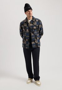 Multi-patterned jacket in shades of blue and beige, featuring two chest pockets, black inner layer, paired with dark trousers and sneakers.