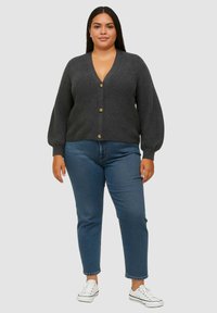 Cardigan lavorato a maglia grigio antracite con scollatura a V, texture a coste e tre bottoni. Abbinato a jeans blu e sneaker bianche.