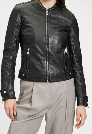 Femme portant une veste en cuir noire ajustée avec des poches zippées et un pantalon beige taille haute sur un fond uni.