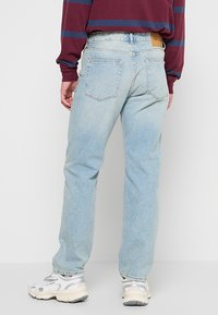 Jeans de mezclilla azul claro con corte recto, cinco bolsillos y un parche de cuero en la parte trasera de la cintura. Llevados con una camisa a rayas color burdeos y zapatillas.