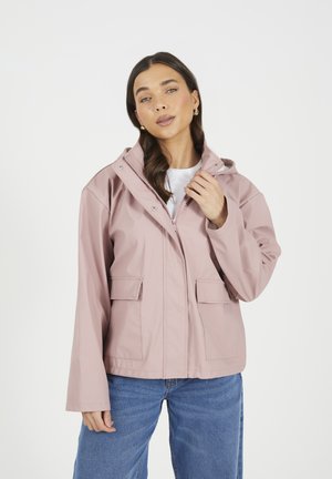 Giacca impermeabile rosa chiaro con cappuccio, chiusura frontale con zip, bottoni a pressione e due tasche laterali. Indossata sopra una maglietta bianca e abbinata a jeans blu.