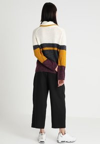 Strickpullover mit hohem Kragen, versehen mit horizontalen Streifen in Creme, Grau, Senf und Burgund. Kombiniert mit schwarzen, weit geschnittenen Hosen.