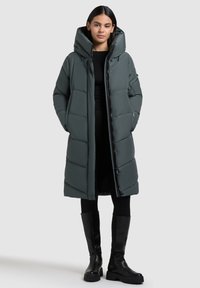 Long, puffy coat in dark green with a high collar and oversized hood. Features a buttoned front and side pockets. Styled with black leggings and boots.