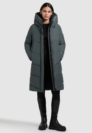 Cappotto lungo e morbido di colore verde scuro con colletto alto e cappuccio oversize. Presenta una chiusura frontale con bottoni e tasche laterali. Abbinato a leggings neri e stivali.