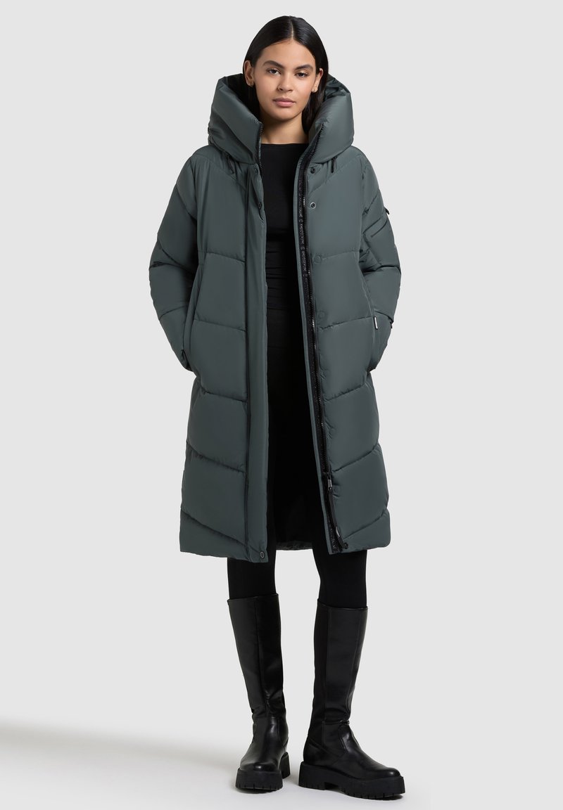 Long, puffy coat in dark green with a high collar and oversized hood. Features a buttoned front and side pockets. Styled with black leggings and boots.