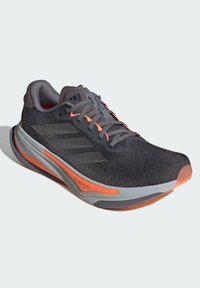 Scarpa da corsa con tomaia in rete nera, accenti grigi, dettagli arancioni e suola texturizzata. Include il logo delle tre strisce e la punta rinforzata.