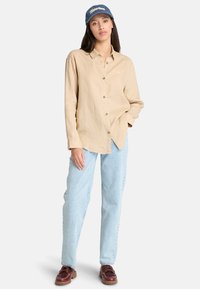 Camicia beige a maniche lunghe con bottoni, abbinata a jeans azzurri e scarpe stringate marroni. Il soggetto indossa un cappellino blu con testo bianco.