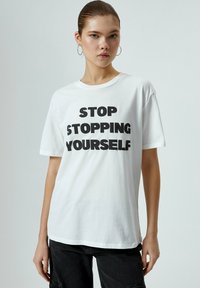 Hvid bomulds t-shirt med et grafisk print, der har stor sort tekst, der står "STOP STOPPING YOURSELF." Klassisk crew neck, afslappet pasform.
