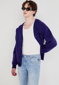 Lilla cardigan med svarte knapper, stripete mansjetter, kombinert med en hvit tanktopp og lyseblå jeans. Inkluderer stilige svarte solbriller.