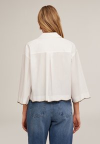 Blusa bianca cropped con maniche corte e larghe, con orlo a scalloped decorato con un bordo di perline. Abbinata a pantaloni in denim blu.