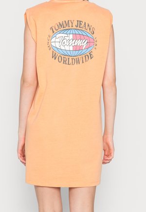 Henkilö, joka pukeutunut oranssiin hihattomaan mekkoon, jossa on takana "Tommy Jeans Worldwide" -logo ja maailman kartta.