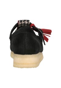 Zwarte suede schoen met een gestructureerde achterkant, met rode accenten en kwastjes, op een lichtgekleurde zool met een geringe vamp hoogte.