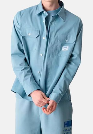 Man die een lichtblauw overhemd met lange mouwen en knopen draagt over een bijpassende blauwe broek, staand met gevouwen handen voor een effen achtergrond.
