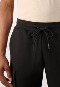 Svart joggebukser laget av mykt stoff med elastisk midjebånd, justerbar snor og sidelommer. Smooth tekstur med minimal detaljering.