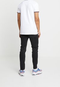 Polo blanco de manga corta con ribetes en rojo y azul marino, combinado con jeans negros y zapatos deportivos multicolores. Fondo liso.