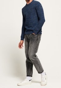 Pull texturé bleu, jeans gris à jambe droite, et baskets blanches avec des accents gris. Le mannequin est de profil, montrant les détails de la tenue.