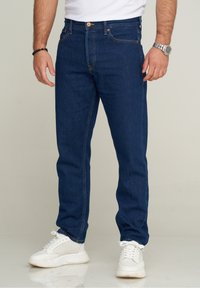 Man som bär mörkblå raka jeans, vita sneakers, vit t-shirt, silverur på höger handled och svart pärlarmband på vänster handled.