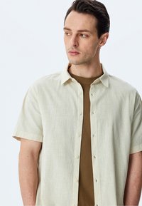Camisa corta de manga corta en un tono beige claro, hecha de una tela texturizada con cierre de botones. Lleva puesta una camiseta de canalé marrón debajo, que presenta un cuello extendido y un bolsillo.