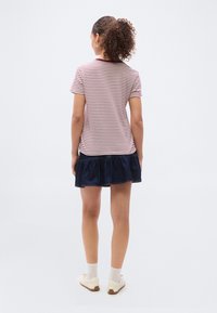 Γυναίκα με σγουρά μαλλιά που φοράει κόκκινη και λευκή ριγέ μπλούζα, σκουρομπλε φούστα με βολάν, λευκές κάλτσες και λευκά αθλητικά παπούτσια, στέκεται με την πλάτη.