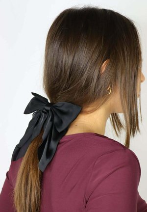 Czarna satynowa kokarda do włosów związana u podstawy kucyka, podkreślająca gładkie, błyszczące włosy. Osoba ma na sobie bordową bluzkę.