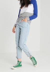 Ljusblå denimjeans med fransade fållar, tillsammans med en långärmad topp i tri-färger med grå, vit och blå sektioner, samt gröna hög-toppsneakers.