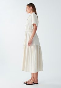 Bežinė, trumpomis rankovėmis maxi suknelė su raukiniu liemenyje, sluoksniuotu apvadu ir mygtukų uždarymu. Derinama su juodais sandalais su auksiniais akcentais.